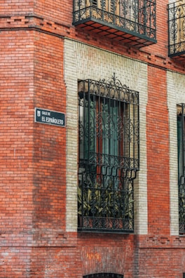 A brick building facade with ornate black wrought iron window grills. A decorative street sign reads 'Calle de El Españoleto.' The windows are bordered by light-colored bricks, contrasting with the red brick exterior.