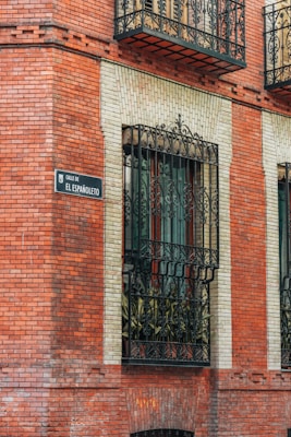 A brick building facade with ornate black wrought iron window grills. A decorative street sign reads 'Calle de El Espa&ntilde;oleto.' The windows are bordered by light-colored bricks, contrasting with the red brick exterior.