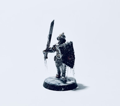 A detailed miniature figurine of a warrior holding a large sword and a shield. The figure is dressed in armor and stands on a small base, with icicles hanging from the shield and sword, suggesting a cold environment.