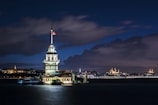 A beautifully illuminated tower stands on a small islet, surrounded by the calm waters of a strait during nighttime. The structure features a prominent Turkish flag flying on top. The background reveals a cityscape adorned with historic architecture, subtly lit, under a partly cloudy night sky.