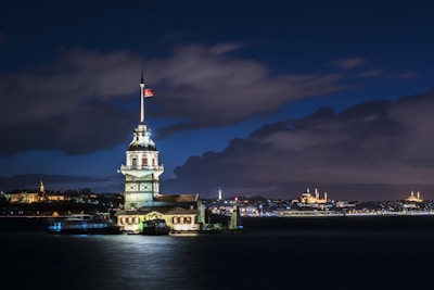 A beautifully illuminated tower stands on a small islet, surrounded by the calm waters of a strait during nighttime. The structure features a prominent Turkish flag flying on top. The background reveals a cityscape adorned with historic architecture, subtly lit, under a partly cloudy night sky.