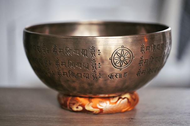 A brass singing bowl adorned with intricate inscriptions and a symbol resembling a dharma wheel. The bowl rests on a decorative cushion with an orange and white pattern. The setting appears to be serene and minimalistic, suggesting a focus on meditation or spiritual practice.