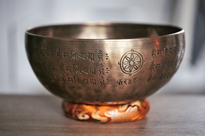 A brass singing bowl adorned with intricate inscriptions and a symbol resembling a dharma wheel. The bowl rests on a decorative cushion with an orange and white pattern. The setting appears to be serene and minimalistic, suggesting a focus on meditation or spiritual practice.