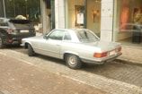 A vintage silver Mercedes-Benz car is parked on a cobblestone street beside a black Range Rover. The vehicles are near an art gallery with large glass windows displaying artwork and sculptures. Autumn leaves are scattered on the ground, adding a rustic touch to the urban setting.