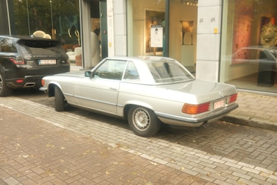 A vintage silver Mercedes-Benz car is parked on a cobblestone street beside a black Range Rover. The vehicles are near an art gallery with large glass windows displaying artwork and sculptures. Autumn leaves are scattered on the ground, adding a rustic touch to the urban setting.