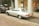 A vintage silver Mercedes-Benz car is parked on a cobblestone street beside a black Range Rover. The vehicles are near an art gallery with large glass windows displaying artwork and sculptures. Autumn leaves are scattered on the ground, adding a rustic touch to the urban setting.