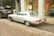 A vintage silver Mercedes-Benz car is parked on a cobblestone street beside a black Range Rover. The vehicles are near an art gallery with large glass windows displaying artwork and sculptures. Autumn leaves are scattered on the ground, adding a rustic touch to the urban setting.