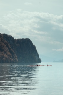 A serene lake with calm waters reflects the surrounding landscape. Dense, forested hills rise on the left side, creating a natural backdrop. The sky is overcast with light cloud cover, and the view includes several kayakers paddling across the water, adding a sense of tranquility and leisure to the scene.
