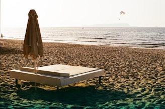 A sandy beach with a large daybed and a closed umbrella in the foreground. The ocean stretches out towards the horizon with gentle waves lapping at the shore. In the distance, there is a parasailer high in the sky. The lighting suggests a setting sun, casting a warm, golden hue over the scene.