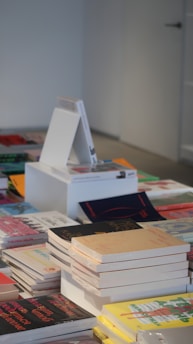 A display of various books stacked and spread out on a table, with a neutral-colored wall in the background. Some books are stacked in piles, while others lay flat. The assortment includes books of different sizes and covers, with a mix of vibrant and subdued colors.