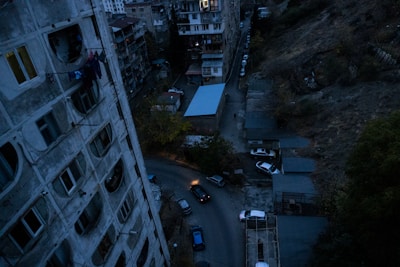 Aerial view of an urban area with tall, worn-looking apartment buildings and a narrow street lined with parked cars. The buildings show signs of aging, with visible marks and laundry hanging outside windows. The lighting is dim, suggesting either early morning or late evening with a somber sky.