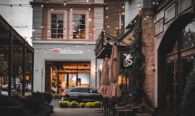 A charming outdoor shopping area illuminated by string lights. The scene features a store with a lululemon sign, with modern architecture and a glass storefront. The pathway is adorned with closed umbrellas and quaint tables and chairs. A dark-colored car is parked in front of the store, and greenery is seen climbing the brick walls.
