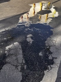 A reflective puddle on an asphalt surface mirroring parts of a structure with white walls and yellow and red vertical elements. The ground shows signs of wear with cracking and discoloration.