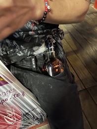 A person is wearing dark clothing and a bracelet with colorful beads on their arm. A bottle, partially concealed, peeks out from the waistband or pocket area. A plastic shopping bag with visible text is held by the person.