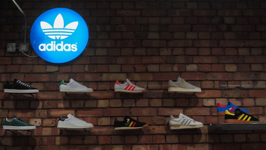 A display of various Adidas sneakers mounted on a brick wall. The blue Adidas logo is illuminated on the top left corner.