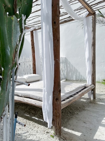 A wooden four-poster outdoor bed with a white mattress and white sheer fabric drapes is set on a sandy surface. The structure features wooden beams and an organic, rustic aesthetic. A large green cactus or succulent plant is partially visible in the foreground, adding to the natural theme. Sunlight casts shadows, creating a bright and airy atmosphere.