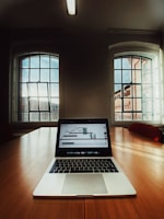 A laptop displaying a stock market graph is placed on a large wooden conference table in a room with tall windows letting in natural light. The room features a minimal design with neutral tones and a ceiling mounted fluorescent light fixture.
