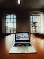 A laptop displaying a stock market graph is placed on a large wooden conference table in a room with tall windows letting in natural light. The room features a minimal design with neutral tones and a ceiling mounted fluorescent light fixture.