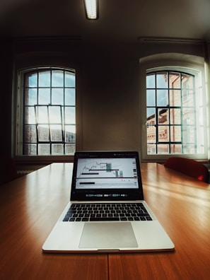 A laptop displaying a stock market graph is placed on a large wooden conference table in a room with tall windows letting in natural light. The room features a minimal design with neutral tones and a ceiling mounted fluorescent light fixture.