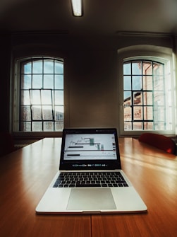 A laptop displaying a stock market graph is placed on a large wooden conference table in a room with tall windows letting in natural light. The room features a minimal design with neutral tones and a ceiling mounted fluorescent light fixture.