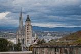 A dramatic skyline view featuring a tall cathedral with ornate spires and a nearby historic tower with a Quebec flag at its peak. In the background, a panoramic landscape of rolling hills and a cloudy sky creates a moody atmosphere. The foreground includes a stone wall covered with patches of grass.