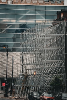 A large metal scaffolding structure is set up in front of a modern building with a glass facade. The scene includes a city street with a few pedestrians walking and cars parked nearby. The architecture features a combination of sleek, contemporary design with some older, decorative elements visible at the top of the building. A lit streetlamp adds a warm touch to the otherwise industrial and urban setting.