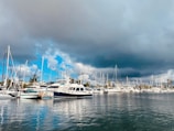 A marina filled with various sailboats and yachts with masts standing tall against a backdrop of a dramatic cloudy sky. The water is calm, reflecting the vessels and the hazy atmosphere. Palm trees are visible in the background, adding a touch of tropical scenery.