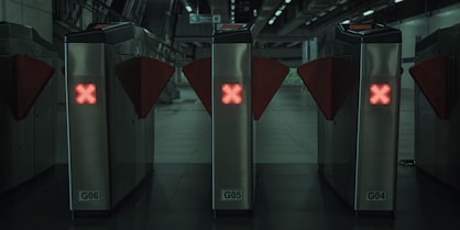 Three metal turnstiles with red glowing X symbols are positioned in a row at a public transit station or subway entrance. The surrounding environment appears dimly lit with a modern and industrial design. Stairs and signage are visible in the background.