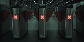 Three metal turnstiles with red glowing X symbols are positioned in a row at a public transit station or subway entrance. The surrounding environment appears dimly lit with a modern and industrial design. Stairs and signage are visible in the background.