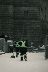 Two individuals in reflective gear walk through a snow-covered area in front of a large stone building. They are equipped for snow removal, each carrying a shovel as snow falls around them.