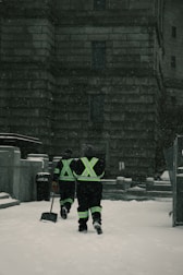 Two individuals in reflective gear walk through a snow-covered area in front of a large stone building. They are equipped for snow removal, each carrying a shovel as snow falls around them.