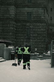Two individuals in reflective gear walk through a snow-covered area in front of a large stone building. They are equipped for snow removal, each carrying a shovel as snow falls around them.