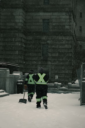 Two individuals in reflective gear walk through a snow-covered area in front of a large stone building. They are equipped for snow removal, each carrying a shovel as snow falls around them.