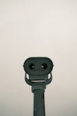 A grey metal telescope positioned against a pale, overcast sky. The telescope is structured with two viewing lenses and a large base, pointing towards the observer. It exhibits signs of wear with slight discoloration and a few marks.