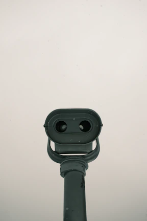 A grey metal telescope positioned against a pale, overcast sky. The telescope is structured with two viewing lenses and a large base, pointing towards the observer. It exhibits signs of wear with slight discoloration and a few marks.