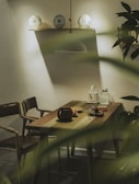 A cozy and intimate dining area features a wooden table with a green table runner. On the table, a dark-colored bowl, a glass, a teapot, and other small items are arranged. Two chairs with a modern design are positioned on each side. Above the table, a pendant light provides soft illumination, and decorative plates are displayed on a shelf. Nearby, green foliage adds a natural touch.