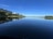 A tranquil lakeside view with a clear blue sky and calm water reflecting the surrounding landscape. High-rise apartment buildings line the left side, bordered by lush green trees. The stillness of the water creates a mirror-like effect, capturing the skyline and greenery.
