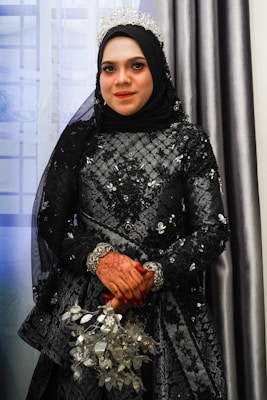 A woman is dressed in an ornate, black embroidered outfit with silver accents and a headpiece, likely a tiara. She is wearing a black hijab. Her hands are decorated with henna, and she is holding a bouquet of silver-toned flowers. The background features curtains and a window with sheer drapes, creating a soft and elegant atmosphere.