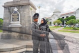 A couple in traditional attire poses near a war memorial. The woman is wearing a hijab and a long black dress, while the man is dressed in a matching traditional outfit. They are embracing each other outside, in an area with historic architecture, greenery, and overcast skies.