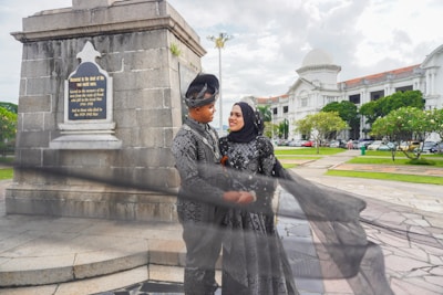 A couple in traditional attire poses near a war memorial. The woman is wearing a hijab and a long black dress, while the man is dressed in a matching traditional outfit. They are embracing each other outside, in an area with historic architecture, greenery, and overcast skies.