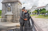 A couple dressed in matching traditional attire poses in front of a monument with a plaque, surrounded by a scenic outdoor setting with a historic building in the background. The woman wears a hijab, and both outfits are intricately patterned. The area features paved walkways and well-maintained greenery.