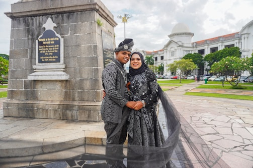 A couple dressed in matching traditional attire poses in front of a monument with a plaque, surrounded by a scenic outdoor setting with a historic building in the background. The woman wears a hijab, and both outfits are intricately patterned. The area features paved walkways and well-maintained greenery.