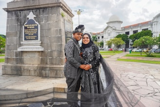 A couple dressed in matching traditional attire poses in front of a monument with a plaque, surrounded by a scenic outdoor setting with a historic building in the background. The woman wears a hijab, and both outfits are intricately patterned. The area features paved walkways and well-maintained greenery.