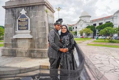 A couple dressed in matching traditional attire poses in front of a monument with a plaque, surrounded by a scenic outdoor setting with a historic building in the background. The woman wears a hijab, and both outfits are intricately patterned. The area features paved walkways and well-maintained greenery.