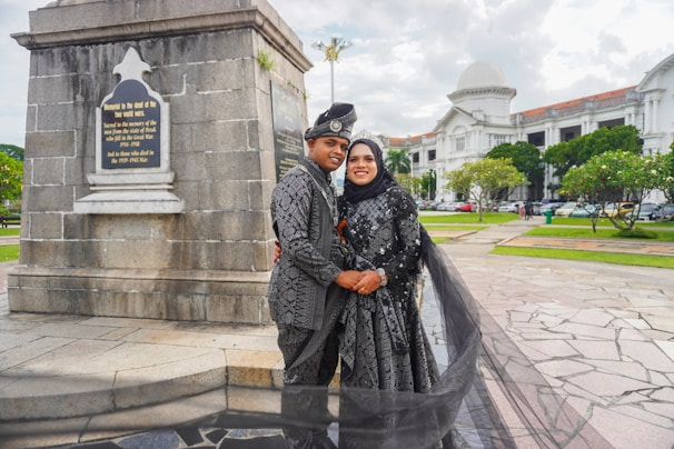 A couple dressed in matching traditional attire poses in front of a monument with a plaque, surrounded by a scenic outdoor setting with a historic building in the background. The woman wears a hijab, and both outfits are intricately patterned. The area features paved walkways and well-maintained greenery.