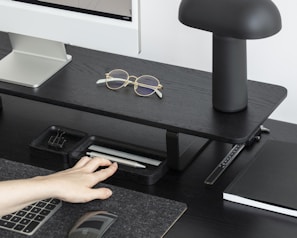 A neatly organized desk setup with a modern aesthetic. The desk features a black wooden stand holding a monitor, glasses placed on top, and a stylish black lamp. Below the stand, there are two trays holding pens and a binder clip. A hand is reaching towards a wireless mouse next to a sleek keyboard. A notebook lies beside the stand.