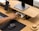 A neatly organized workspace featuring a wooden desk with a monitor on a stand, a wireless mouse on a dark mouse pad, and a hand reaching for a game controller stored underneath the monitor stand. A smartphone is positioned on a stand next to the monitor.