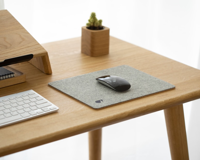 A minimalist desk setup features a light wooden desk with a felt mouse pad and a sleek black computer mouse. A modern, compact keyboard sits nearby, and there is a small wooden stand holding notebooks. In the background, a small potted succulent adds a touch of greenery.