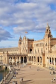 A grand architectural structure with ornate details and numerous arches extends into a large plaza. People are scattered throughout the open area, enjoying the sunny day. A Spanish flag waves atop one of the building's towers. The sky is partly cloudy, creating a pleasant atmosphere.