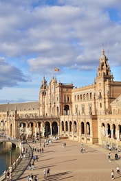 A grand architectural structure with ornate details and numerous arches extends into a large plaza. People are scattered throughout the open area, enjoying the sunny day. A Spanish flag waves atop one of the building's towers. The sky is partly cloudy, creating a pleasant atmosphere.